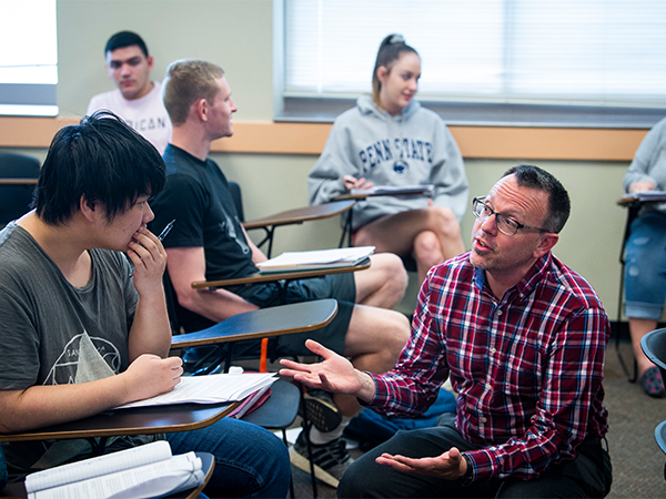 An instructor kneels down in front of a student in a classroom and speaks with that individual.